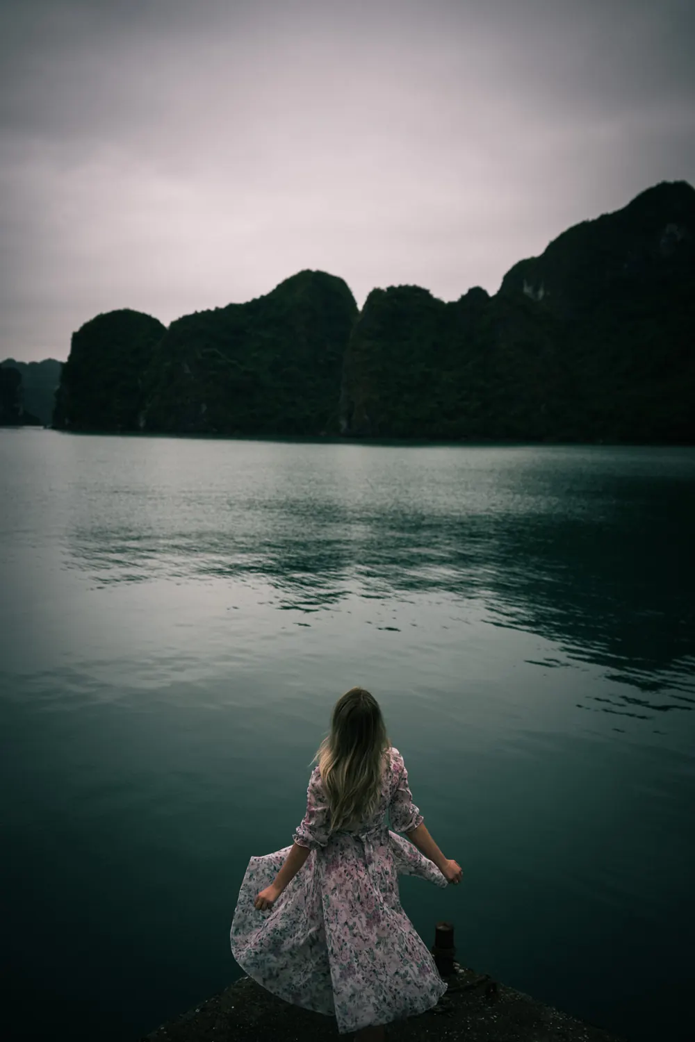 Perempuan bergaun di dermaga, menghadap teluk dan bukit karst saat mendung, dengan preset Ricoh Positive